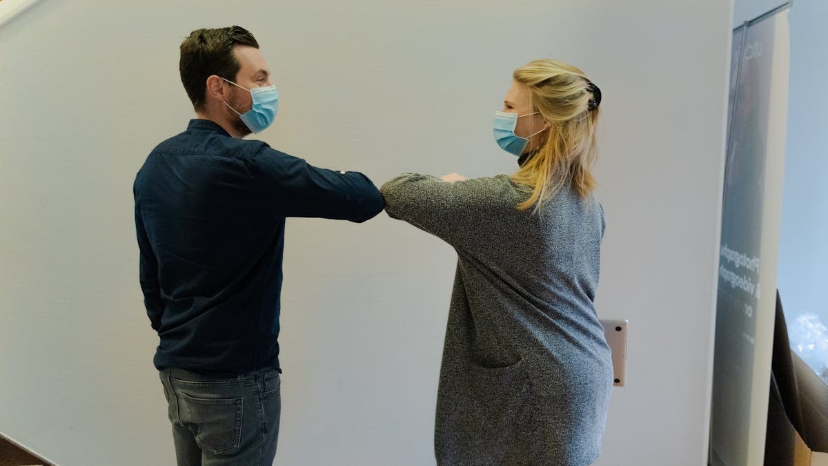 Two colleagues greet with an elbow bump while wearing face masks indoors.
